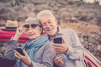 Cheerful old senior couple use phones together outdoor with red car in background - concept of travel mature people and modern