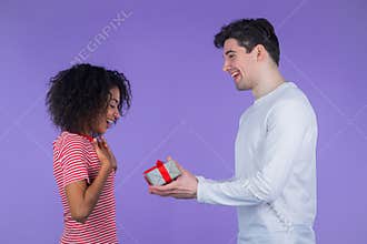 Young interracial couple on purple studio background. White man gives gift with red bow to his african american wife or