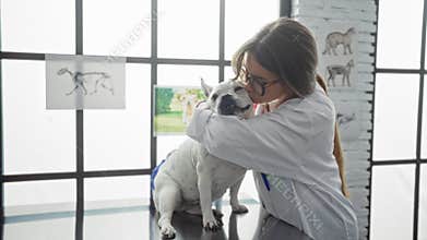 A blonde woman in a veterinary clinic kisses a white dog while standing indoors with medical diagrams in the background
