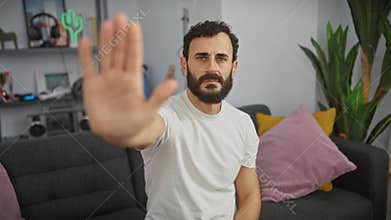 A serious bearded man gestures a stop sign with his hand in a modern living room setting, portraying personal boundaries or