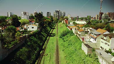 Flying over empty railroad track, rail way in green grass among city buildings in summer, sunny day. Buenos Aires