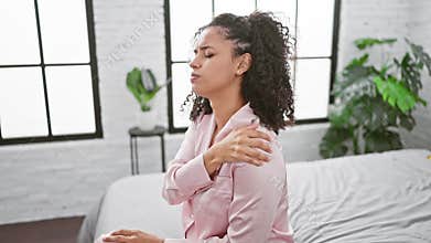 Young, beautiful hispanic woman in morning bed, suffering severe shoulder pain, unhappily clutching aching area in bedroom