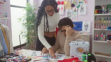 A woman and a man collaborate in a tailoring atelier among sewing machine, fabrics, sketches, and thread