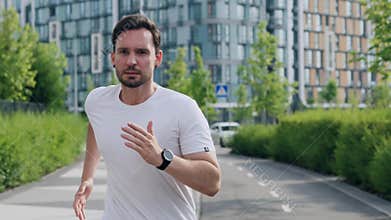 A young positive sportive man does morning jogging on the sidewalk, wipes forehead from sweat, checks the time on the