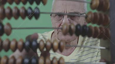Old man wearing glasses uses abacus with brown and black beads. He is dressed in yellow striped sweater and focused on