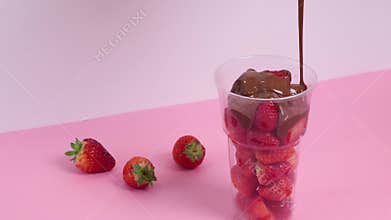 Process of pouring strawberry berries with melted liquid chocolate in cup. Sweet food, dessert on pink background.