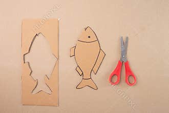 a piece of cardboard with a fish shape cut out of it, step in creative process, red handle scissors, top view, beige