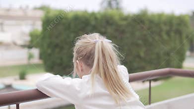 A little girl in pajamas stands on the balcony in the morning and looks at the pool