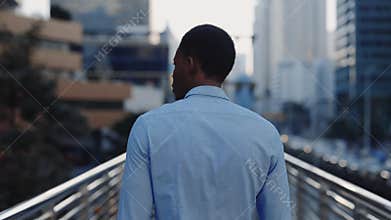 Back View of Young Successful African American Man in Shirt Walking Across Bridge in Business District. The Back of