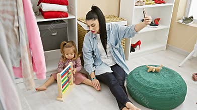 A woman takes a selfie while her daughter plays with an abacus in a cozy dressing room filled with shoes and clothes