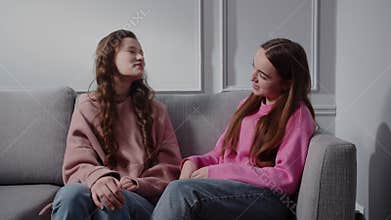 Two smiling positive charming teenagers girls sit on the sofa in the room and talk. Girlfriends or sisters have