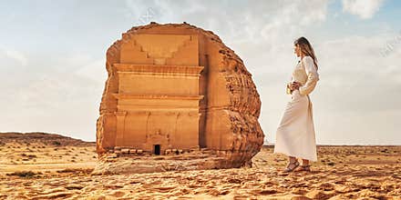 Young Caucasian woman in long white dress posing in front Tomb Lihyan Son of Kuza or Qasr al-Farid at Hegra, Saudia Arabia, sandy