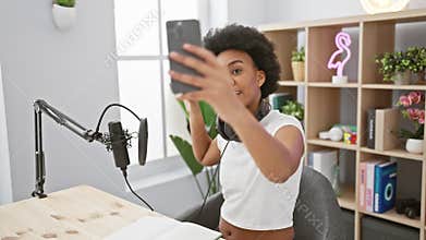 An african-american woman takes a selfie in a modern radio studio with professional podcasting equipment