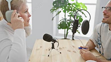 A man and woman engage in podcasting indoors, sitting at a wooden table with microphones and headphones in a light-filled studio