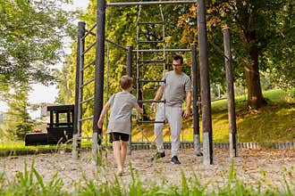 A father and his young son engage in joyful playtime at a playground surrounded by vibrant greenery. They share laughter and