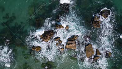 Aerial view of waves crashing over coastal rocks on a turquoise sea, with a scenic sandy beach and forested cliffs in the