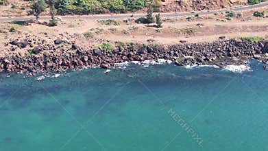 Aerial view of a scenic coastal road running along a rugged, rocky shore with clear turquoise ocean water in Maharashtra, India