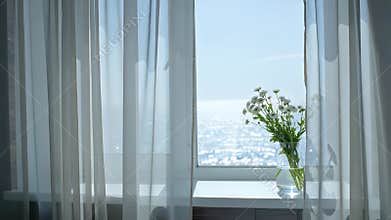 Bright airy windowsill framed by sheer curtains overlooking the sparkling sea with a vase of flowers