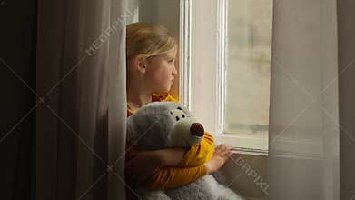 Frustrated lonely Caucasian girl child kid orphan daughter looking out window holding teddy bear bored victim innocence