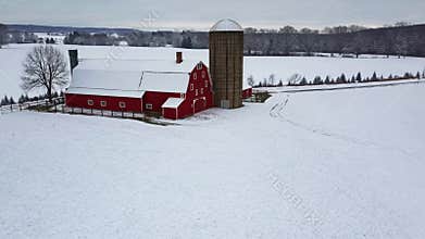 Snow blankets a red barn and silo in a tranquil winter landscape with rolling hills and trees