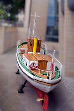 Model of a vintage tugboat displayed on land near a modern building in an urban area