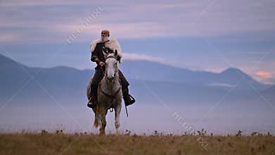 Elderly Viking warrior in fur cloak riding gray horse across open field at sunrise, mountains and distant cross behind