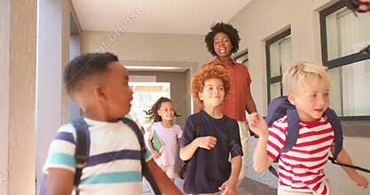 Diverse kids with backpacks exiting walkway toward yard, speeding with teacher holding tablet