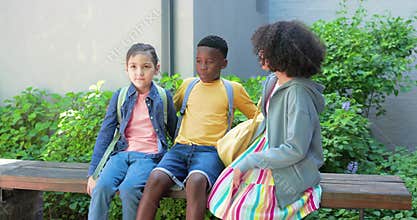 Diverse kids exiting walkway with backpacks sitting on bench while chatting and playing hand game