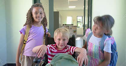 Three school-age children arriving at school doorway, posing around wheelchair and fixing backpacks