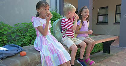 Three children sitting on brick ledge, sunlight causing boy rubbing eyes, gesturing, backpacks near