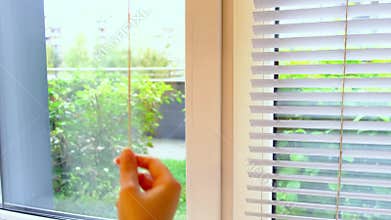 Side view shows a hand adjusting white window blinds, soft daylight and blurred movement creating a clean minimal composition