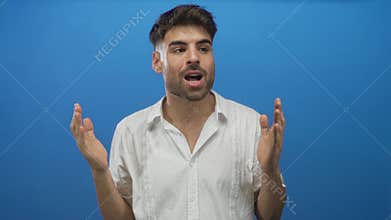 Young hispanic man with open mouth, raised hands and surprised expression in bright blue studio; surprise amazement