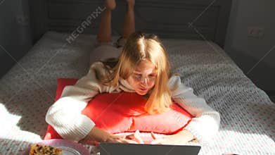 Casual young woman lying in bed, writing a blog post or article on her laptop