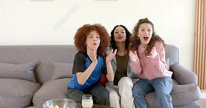 Diverse female friends reacting to basketball, joining, cheering on sofa with chips and lemonade