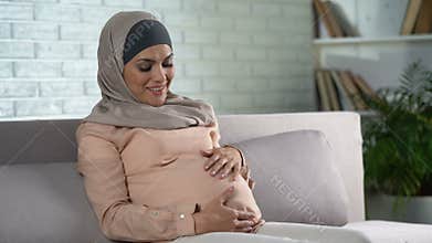 Happy pregnant woman in hijab touching belly gently, resting home, prenatal care