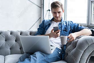 Young man using his smartphone for online banking - sitting on sofa with laptop on leap.