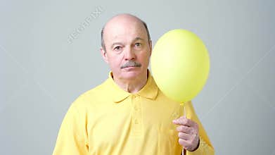 A sad senior man in a yellow shirt is holding a small balloon in his hands.