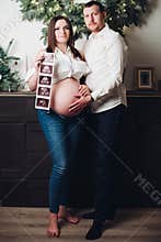 Pregnant woman posing with man, holding ultrasound pictures.