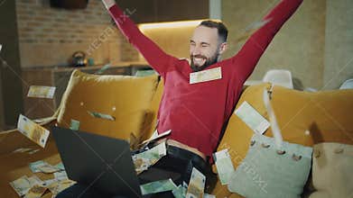 Happy bearded man is working at home with laptop and making a successful deal or win big money. Money falls from above