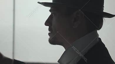 Portrait of handsome confident man putting a hat on the head in front of big window. The head of the mafia preparing for