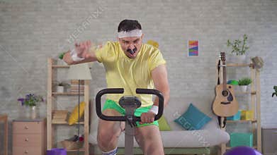 Funny energetic athlete of the 80`s with a mustache is engaged on a exercise bike and pours water from a bottle on his
