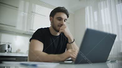 Smiling man using laptop computer at kitchen table. Young business man smiling