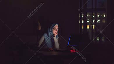 Muslim girl with glasses and a hijab with notebook in the dark search on the Internet at home