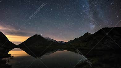 Milky Way Time lapse starry sky beyond snowcapped mountain ridge, reflected on idyllic apine lake