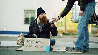 Young man helps to homeless person and giving him some food while beggar drink alcohol and sit near shopping cart at