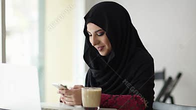 Portrait of young attractive smiling muslim woman in hijab sitting in the cafe and typing a message on her mobile phone