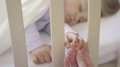 The parent holds the hand of a small child sleeping in a baby crib. Happy family and her newborn baby together. The