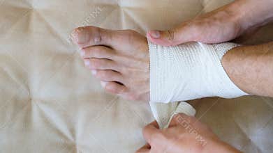 First aid man applying bandage for injured foot and ankle sitting on couch at home.