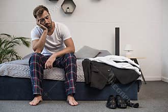 Young sleepy man in home clothes sitting on bed after waking up