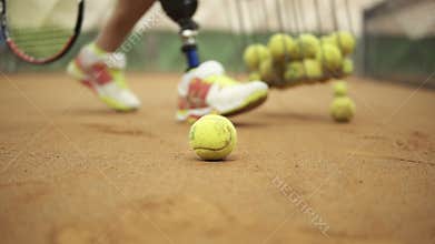 Closeup footage of female legs in sports sneakers and prosthesis on her leg picking up tennis balls from the tennis